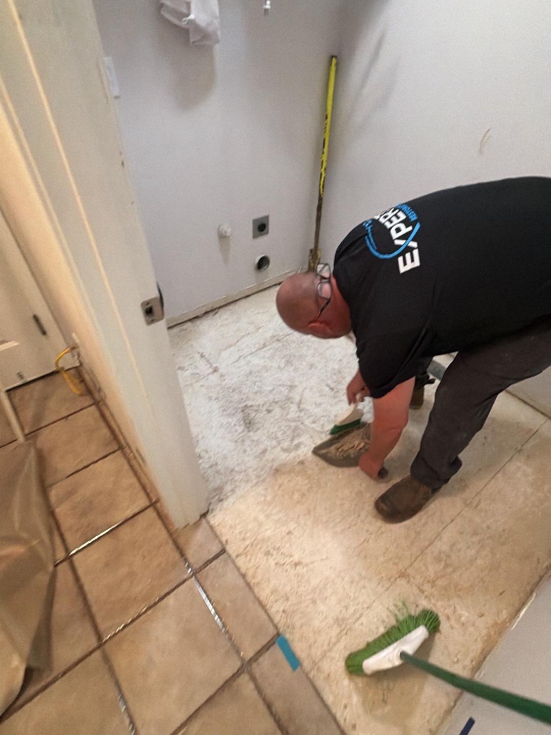 Expert Restoration team member in safety glasses and branded shirt sweeping debris from an exposed laundry room subfloor after controlled demolition