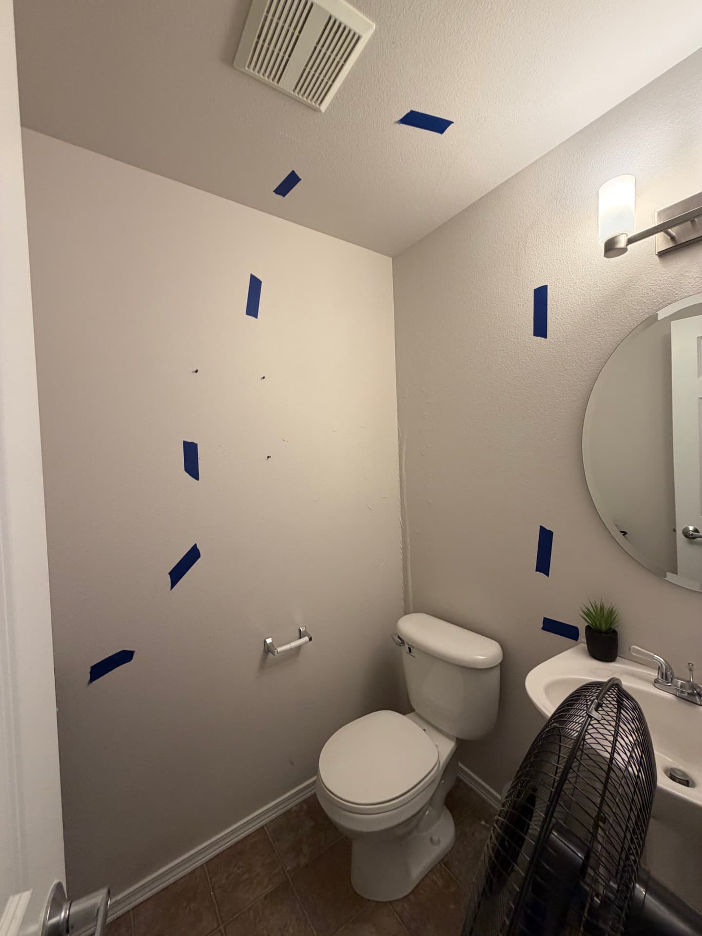 Bathroom with an air mover on the floor and blue tape marking damaged drywall behind the toilet